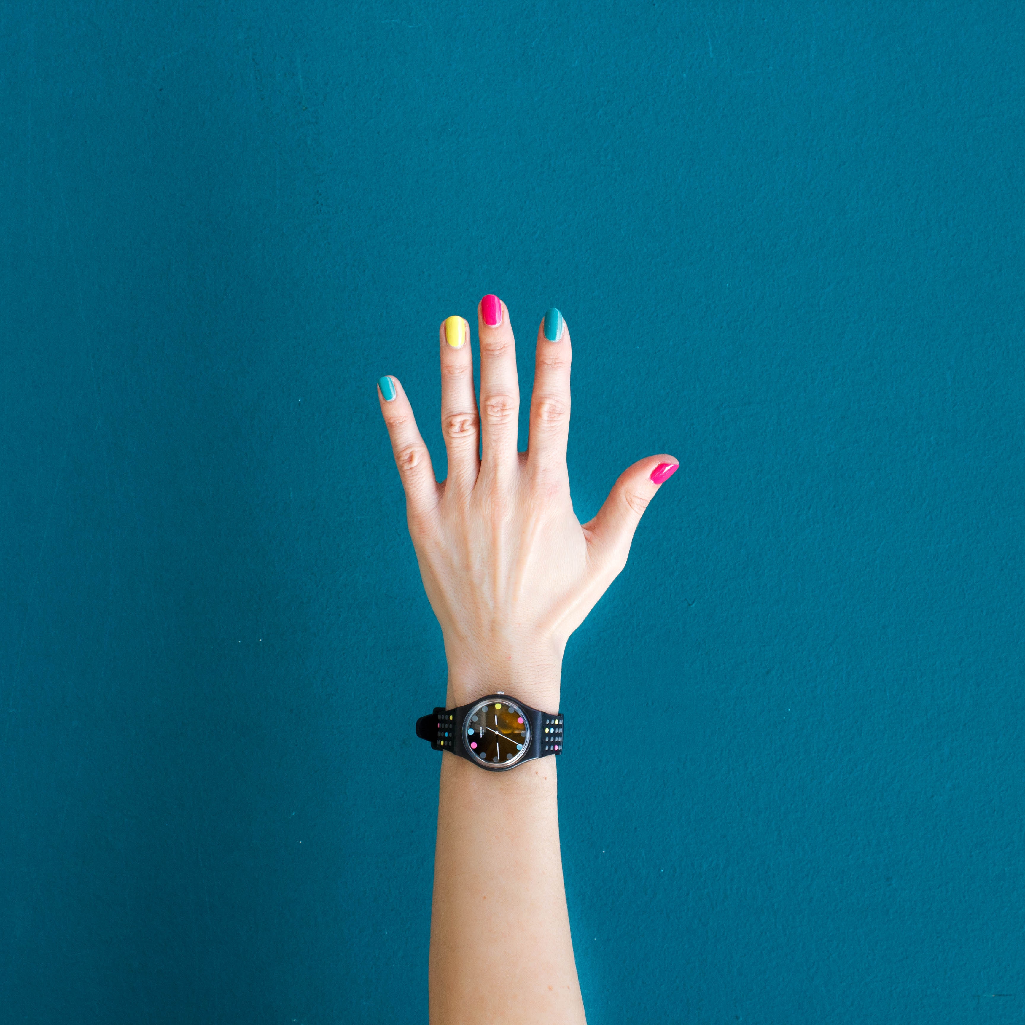 A hand raised in front of a blue background.  It is wearing a watch, and has colorfully painted nails.  