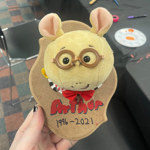 Hand holding a faux-taxidermy trophy composed of a stuffed animal "Arthur" head from the TV Series Arthur. The dates 1996-2021 are written below the mounted head.