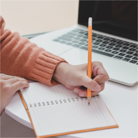 Photo: A hand writes with a pencil in a lined notebook in front of an open laptop. 