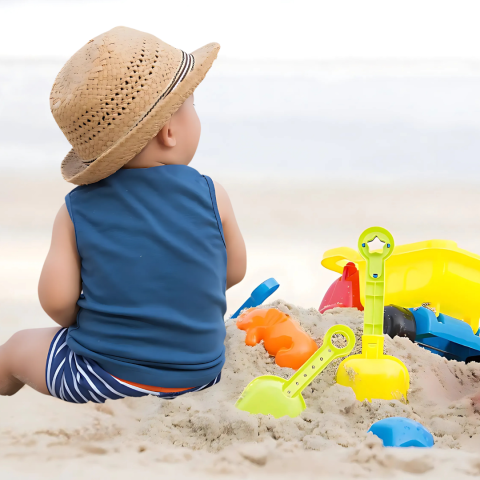a baby facing away from the camera in a blue shirt and tan straw hat sits on a white sandy beach with a mound of sand and colorful beach toys next to them