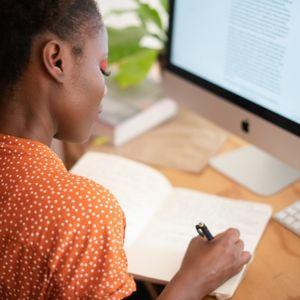 woman writes in notebook in front of computer