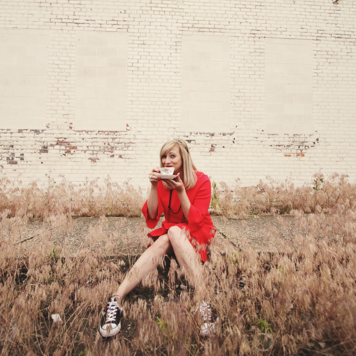 performer Melissa Behring sits on a sidewalk among tall grasses sipping from a cup of tea