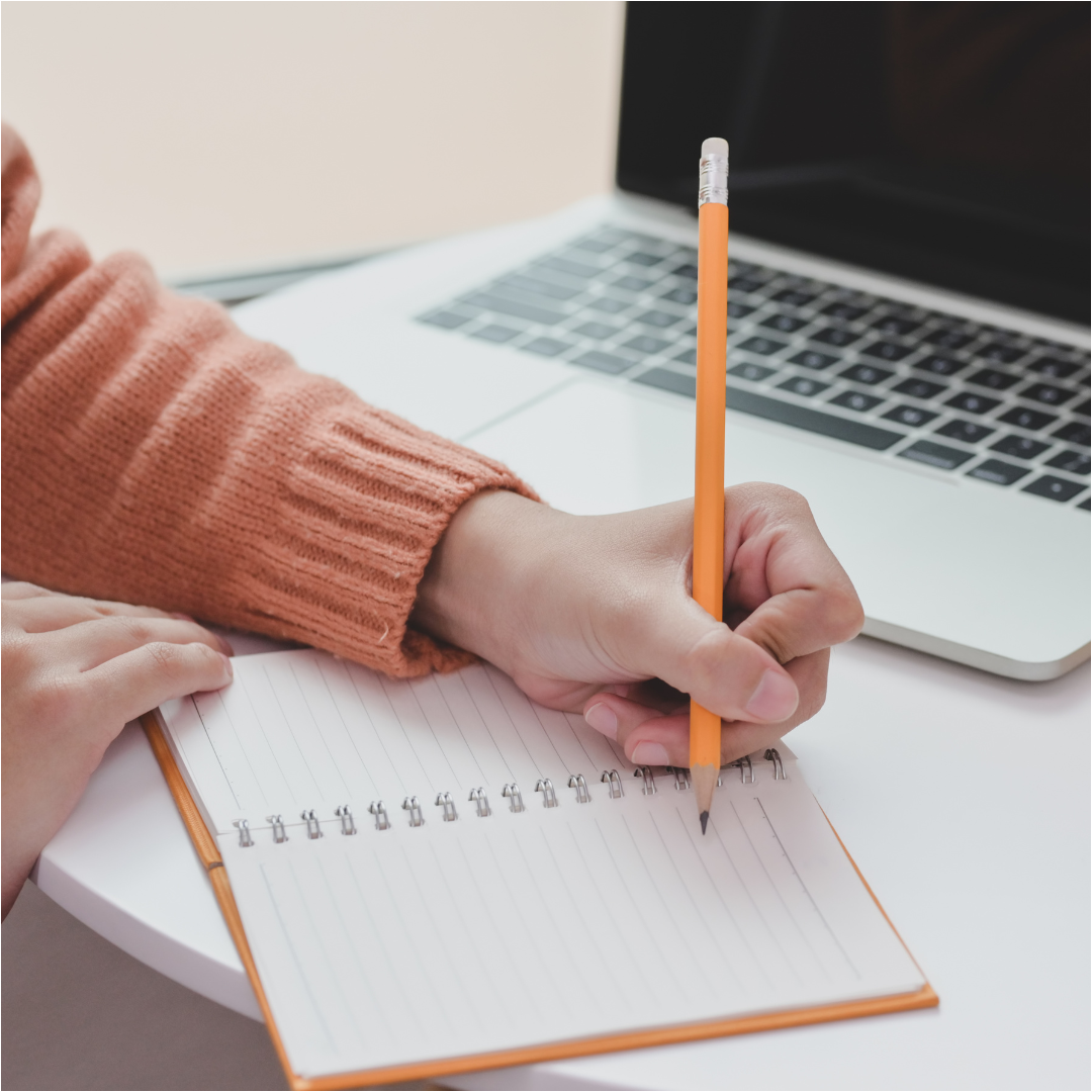 Photo: A hand writes with a pencil in a lined notebook in front of an open laptop. 