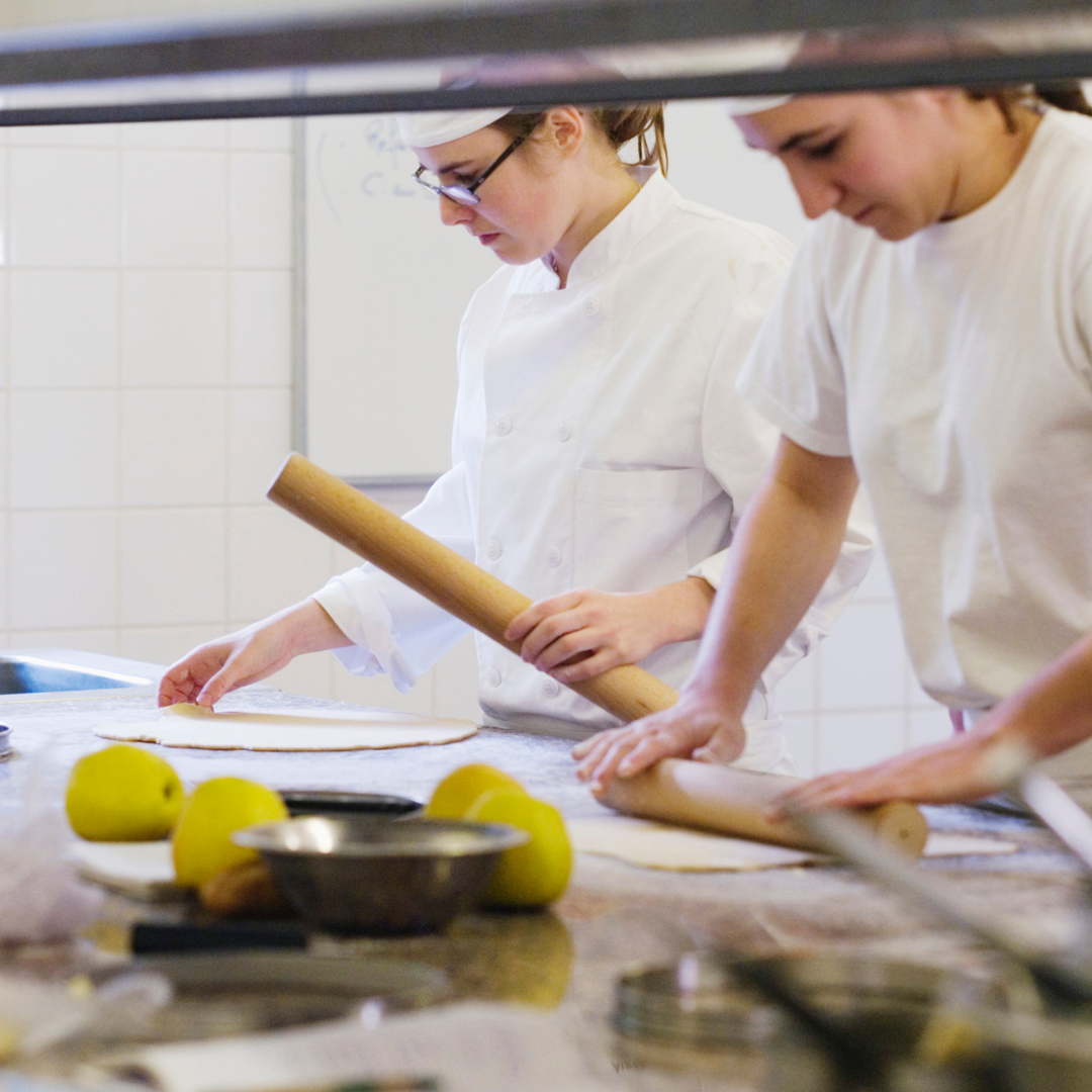 Two people in chef's coats rolling out dough. 